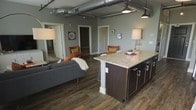 This stylish loft-style residence blends industrial character with modern comfort in a thoughtfully designed single-floor layout. The open-concept living and kitchen area serves as the heart of the space, where exposed metallic ductwork along the ceiling adds an authentic industrial edge, while rich dark hardwood-style flooring grounds the design with warmth and continuity throughout.

The kitchen is a standout feature, anchored by a generous island topped with light granite countertops and finished with sleek dark cabinetry accented by modern hardware. The island doubles as a casual dining and gathering spot, seamlessly connecting the kitchen to the living area. Pendant lighting above reinforces the industrial aesthetic while providing practical illumination.

The living area is open and inviting, with ample room for comfortable furnishings. Soft, neutral wall tones in a cool sage-gray palette complement the overall design and create a calm, cohesive ambiance. Multiple doorways visible from the main living space suggest well-defined private rooms, offering a balance of open communal areas and separated personal spaces.

Track and pendant lighting are thoughtfully placed throughout, ensuring the space is well-lit while contributing to its modern, urban character. Overall, this residence offers a dynamic and functional living environment with a distinctive loft-inspired personality.