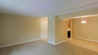 This single-floor space presents an open and airy layout designed for comfortable everyday living. The main living area is finished with wall-to-wall carpeting in a neutral beige tone, complementing the warm, light tan walls and crisp white trim and baseboards throughout. A textured white ceiling adds subtle character to the space, while a centrally placed HVAC vent ensures consistent climate control.

A structural column divides the living area, creating a natural sense of separation between zones without fully enclosing the space. The front entry features a classic six-panel white door, providing a clean and welcoming point of arrival. Crown molding detail is visible along the ceiling near the entryway area, adding a refined architectural touch.

Adjacent to the main living space, a passthrough opening leads into the kitchen area, where wooden cabinetry in a warm finish is visible alongside what appears to be a dark-toned appliance. The transition from carpeted living space to a hard-surface kitchen floor is subtly distinguishable, reinforcing the functional separation of areas. Overall, the space conveys a straightforward, well-maintained interior with a neutral palette suited to a variety of furnishing styles.