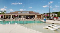 Located at 9921 Horizon Overlook Dr in Raleigh, NC, this single-level outdoor amenity complex offers a resort-style experience centered around a generously sized swimming pool with a shallow wading ledge, in-water lounge chairs, and decorative water features. The pool deck is surrounded by ample seating areas outfitted with lounge chairs and colorful umbrellas, ideal for relaxing in the Carolina sun.

A covered pavilion runs along one side of the pool, featuring a coffered ceiling with ceiling fans for year-round comfort. This shaded retreat is furnished with upholstered wood-framed lounge seating, low coffee tables, and a wall-mounted television — creating an inviting outdoor living room atmosphere. A built-in grill station and pass-through windows further enhance the entertainment potential of the space.

The covered porch area also includes a ping-pong table and vibrant accent seating in bold pink, accessible through bold blue French doors. Brick and board-and-batten siding give the clubhouse structure a classic yet contemporary aesthetic. A pergola area at one end of the deck provides additional shaded seating. Open views of the surrounding community and lush treeline add a scenic backdrop to the entire outdoor experience.