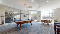 Located at 1401 King in Bridgeport, Connecticut, this contemporary game room is designed as a well-appointed recreational amenity space. The room is finished with patterned blue-toned carpeting throughout, lending a cohesive, modern aesthetic that ties the space together.

At the center of the room sits a solid wood billiards table with a blue felt surface, illuminated by a sleek pendant light fixture suspended from a coffered ceiling detail above. Recessed lighting supplements the abundant natural light that pours in through floor-to-ceiling windows along the exterior walls, offering views of the surrounding streetscape and greenery.

A warm wood-paneled accent wall serves as a focal point on one side of the room, anchoring a wall-mounted flat-screen television. Glass partition walls and sliding doors provide separation from adjacent spaces while maintaining an open, airy feel throughout.

In a sun-drenched corner, a dedicated card and poker table surrounded by upholstered mid-century modern chairs creates an intimate seating area. Along one wall, a wall-mounted counter-height shelf lined with blue-cushioned bar stools offers additional casual seating beneath a curated gallery wall featuring framed artwork, maps, and game-themed prints, adding personality and a playful character to the overall design.