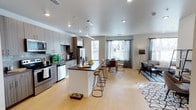This single-floor residence showcases a thoughtfully designed open-concept layout that seamlessly integrates the kitchen, dining, and living areas into one cohesive, light-filled space. The contemporary kitchen is appointed with stainless steel appliances, including a refrigerator, dishwasher, and range, complemented by modern flat-front cabinetry in a warm grey finish. A central island with a dark countertop and bar seating serves as both a functional workspace and a natural gathering point for casual dining. Pendant lighting above the island adds a stylish focal point, while recessed ceiling lights illuminate the entire open floor plan. Light-toned hardwood flooring flows throughout, lending warmth and visual continuity across the space. Large windows dressed with patterned curtains invite abundant natural light and provide pleasant outdoor views, creating an airy and welcoming ambiance. The dining area comfortably accommodates a full table and chairs, while the adjacent living area offers a relaxed setting for lounging. The overall design balances clean, modern aesthetics with functional versatility, making this a well-appointed and inviting space suited for both everyday living and entertaining.