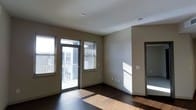 This single-floor space is characterized by its clean, contemporary design and practical layout. The main living area features rich, dark hardwood flooring that contrasts with the neutral, light-toned walls, creating a balanced and modern aesthetic. A glass-paneled door leads directly to an outdoor balcony, allowing natural light to flood the interior while providing access to exterior views of the surrounding urban environment. An adjacent window with blinds further enhances the natural light in the space. The smooth, white ceiling contributes to an open and airy atmosphere. A doorway visible along the right wall connects to an additional room, suggesting a functional layout with clear separation of living spaces. White baseboards frame the perimeter, adding a crisp finishing detail to the interior. Electrical outlets are conveniently positioned along the walls. Overall, the space presents a straightforward, well-lit environment with durable finishes and thoughtful design elements suited for everyday living.