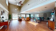 Located at 200 Branchwood Dr in Goose Creek, South Carolina, this single-floor space offers an open, airy layout that seamlessly integrates a living room and dining area into one expansive, well-appointed environment. The most striking architectural feature is the dramatic vaulted ceiling accented by shiplap wood paneling, which soars overhead in the main living zone and is complemented by dual ceiling fans. Rich, warm hardwood flooring runs throughout the entire space, adding continuity and a sense of inviting comfort.

The living area is generously proportioned, furnished with comfortable seating and bathed in natural light streaming through large sliding glass doors and multiple windows that offer direct views of an outdoor pool and deck. A reception-style console table near the entrance provides a functional focal point, with wall art adding character to the neutral taupe walls.

The dining area accommodates multiple round glass-top tables with stylish seating, making it well-suited for group gatherings. A floor-to-ceiling stone fireplace anchors this zone, with a wall-mounted television above, creating both a cozy atmosphere and a practical entertainment feature. Crown molding and clean white trim throughout lend a polished, finished look to the overall space.