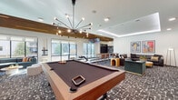 This contemporary entertainment lounge is thoughtfully designed as a premier social and recreational destination. The open, single-floor layout encompasses a rich variety of amenities, all unified by a bold geometric-patterned carpet that anchors the space's modern aesthetic.

At the heart of the room stands a full-size pool table, illuminated by a striking mid-century-inspired sputnik chandelier with multi-directional arms and exposed bulbs. Adjacent to it, a dedicated bowling lane is paired with a long communal high-top table and bar-height seating, with overhead monitors enhancing the experience. Large floor-to-ceiling windows along one wall flood the area with natural light and provide views of the surrounding outdoor landscape.

A vibrant game zone features bold red-patterned wallcovering, a classic arcade cabinet, a jukebox, and additional wall-mounted screens beside a media credenza, creating an energetic retro-modern atmosphere. Plush sectional seating with curated artwork nearby offers a comfortable retreat for spectators and socializers alike.

Glass-partitioned walls with black steel frames define more intimate areas within the lounge, adding an architectural sophistication while maintaining visual openness. Warm wood slat ceiling detailing in select zones adds texture and warmth, completing this well-appointed, amenity-rich entertainment space.