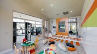 The Fitness Center Kids Room is a thoughtfully designed, child-friendly space that keeps young ones entertained while parents make use of the adjacent fitness center. The two areas are connected via a glass partition wall and a direct door, maintaining visual supervision between the spaces.

The kids' room features a bright, energetic aesthetic defined by bold orange, green, and coral horizontal stripes along the upper walls, complemented by white subway tile wainscoting below. A striking orange accent wall frames a built-in media center with a flat-screen TV, flanked by a low white cubby shelf ideal for storing toys and books. An orange and grey striped area rug anchors the central floor space, which is dotted with soft, stone-shaped floor cushions perfect for lounging.

A dedicated activity zone features child-sized round tables and animal-themed chairs, along with colorful books and games to encourage creativity and learning. Large floor-to-ceiling windows flood the room with natural light and offer open views of the landscaped courtyard and surrounding buildings, creating a bright and welcoming atmosphere. Recessed lighting provides additional illumination throughout. The overall design balances playful energy with practical functionality, making it an engaging and safe environment for children of all ages.