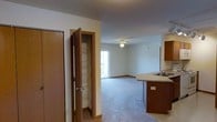 This single-floor residence offers a practical and comfortable open-concept layout that seamlessly connects the kitchen and living areas. The kitchen is outfitted with warm wood-toned cabinetry, a countertop with an integrated sink, and a breakfast bar that provides a natural transition into the main living space. A tall built-in pantry with wire shelving sits conveniently adjacent to the kitchen, offering generous and well-organized storage. The living area features plush wall-to-wall carpeting that adds warmth and comfort underfoot, complemented by neutral-toned walls that create a clean, versatile backdrop. A ceiling fan with a light fixture is centrally positioned overhead, providing both air circulation and ambient lighting. At the far end of the living area, a sliding glass door or wide window opens to a balcony, allowing natural light to fill the space and providing a view of the outdoors. The transition between the kitchen's hard flooring and the carpeted living area is marked by clean wood-toned baseboards and trim that carry throughout the space, tying the design together with a cohesive, warm aesthetic. The overall layout is efficient, bright, and well-suited for everyday living.