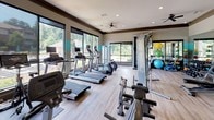 Located at 110 Town Loop in Mooresville, NC, this single-story fitness center is designed to support a wide range of workout routines in a spacious, open layout. The interior features light hardwood-style flooring throughout, recessed lighting, and ceiling fans that contribute to a comfortable exercise environment.

Large windows line the perimeter walls, allowing ample natural light to fill the space and offering views of surrounding greenery. A teal accent stripe runs along the walls, adding a vibrant, modern touch to the neutral color palette. Full-length mirrors on one wall enhance the sense of openness while serving as a practical training aid.

Cardio equipment includes Life Fitness treadmills, elliptical trainers, stationary bikes, and a recumbent bike, many equipped with individual monitors. A Treadwall motorized climbing wall provides a unique functional fitness option. The strength training area offers a multi-station cable machine, a tiered dumbbell rack with a comprehensive weight range, and an extensive collection of medicine balls and stability balls on dedicated storage racks. A wall-mounted television and designated stretching areas round out the facility's amenities, making it suitable for diverse fitness goals.