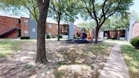 This outdoor recreational space is a well-appointed playground situated within the grounds of a multi-story brick residential community. The area is shaded by a generous canopy of large, mature trees, creating a cool and inviting environment for outdoor play. The centerpiece of the space is a multi-level play structure finished in bold red, yellow, and blue tones, featuring an impressive spiral slide, a straight slide, a climbing wall with hand grips, monkey rings, and a series of ladders and staircases that connect the various platforms. A secondary play structure offers additional climbing bars and activity elements, catering to a range of ages and activity levels. The ground beneath the equipment is covered in dark rubber mulch, providing a safe and impact-absorbing surface. The surrounding open lawn area offers additional space for free play and recreation. The brick residential buildings frame the playground on multiple sides, and exterior staircases are visible in the background, reflecting the functional design of the broader community. The combination of mature tree coverage, colorful equipment, and thoughtfully maintained grounds makes this playground a welcoming and practical outdoor amenity for residents.
