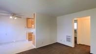 This single-floor space showcases a practical, open-concept layout that connects the living area to the kitchen. The living area is finished with dark wall-to-wall carpeting, providing a warm underfoot feel, while white walls and a textured ceiling contribute to a clean, neutral aesthetic throughout. A wall-mounted floor vent is positioned in the living area, indicating a built-in heating system. A partial wall separates the living space from the kitchen, creating a sense of division while maintaining an open feel. The kitchen features light wood cabinetry, white countertops, and a white range, offering a cohesive and functional cooking space with light-toned flooring. An additional doorway visible from the living area suggests access to a separate room or hallway, adding to the overall flow of the floor plan. The textured ceilings are consistent throughout, tying the spaces together. Overall, the layout is straightforward and efficient, well-suited for everyday living.