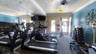 This single-story fitness room is designed to support a well-rounded exercise routine in a compact yet functional setting. The space is outfitted with multiple cardio machines, including treadmills and an elliptical trainer, positioned to allow comfortable use without overcrowding. A multi-functional cable resistance station anchors one side of the room, providing options for strength training exercises. Mirrored walls along one side visually expand the room and allow users to monitor their form during workouts. The flooring is finished in a durable, dark carpet suitable for gym use. Crown molding and a textured ceiling add a degree of architectural refinement, while a ceiling-mounted fan aids in ventilation and comfort during exercise. A wall-mounted flat-screen television offers entertainment during cardio sessions. Natural light filters in through multiple windows and a glass-paneled door, contributing to an open and energizing atmosphere. An accent wall painted in a cool teal tone adds a subtle pop of color, contrasting with the neutral beige walls throughout the remainder of the room.