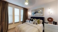 This bedroom is thoughtfully designed to balance comfort and style within a single-floor layout. The room centers around a substantial upholstered bed with a tufted black headboard, dressed in neutral, striped bedding and accented with an assortment of decorative pillows in olive green, black, and patterned tones. Above the headboard, a gold-framed artwork featuring a bold bird motif serves as a striking focal point, adding an artistic touch to the space.

Natural light enters through two windows fitted with white horizontal blinds, complemented by floor-length brown curtains that add warmth and depth to the room. A sleek wall-mounted sconce with dual globe lights provides soft ambient lighting alongside the ceiling fan fitted with an integrated light fixture, ensuring both form and function.

The walls are finished in a soft, neutral tone, contributing to the room's calm and inviting atmosphere. White furniture pieces, including a low dresser beneath the windows, offer practical storage while maintaining the room's clean aesthetic. The overall ambiance is modern yet cozy, with a carefully curated mix of textures and colors.