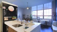 This single-floor space showcases a thoughtfully designed open-plan layout that seamlessly integrates the kitchen, dining, and living areas into one cohesive environment. The kitchen is anchored by a generous white quartz island with an undermount sink and modern brushed-nickel faucet, providing ample workspace and a natural gathering point. Clean white cabinetry and minimalist finishes contribute to the contemporary aesthetic throughout.

A standout feature is the expansive grid-style windows along one wall, flooding the interior with abundant natural light and offering views of the surrounding urban streetscape. The bright, airy atmosphere is further enhanced by white walls and light-toned surfaces. A ceiling fan with a pendant-style mount adds both functionality and a subtle industrial touch overhead.

The space also incorporates a dedicated desk nook positioned near the windows, making it suitable for work or study. A chalkboard accent wall near the kitchen adds a playful, functional element, while open shelving and greenery introduce warmth and personality. Overall, the space balances modern design with a welcoming, lived-in feel.