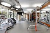 Located at 4501 Ford Ave in Alexandria, VA, this well-appointed fitness center offers a comprehensive workout environment designed with both functionality and aesthetics in mind. The single-floor space is generously sized, providing ample room for a full range of exercise equipment without feeling crowded.

The facility is characterized by its striking industrial-modern design, featuring an exposed ceiling with visible ductwork and oversized circular pendant lights that create a bright, energizing atmosphere. A dramatic wall of floor-to-ceiling windows lines one side of the gym, allowing abundant natural light to pour in while offering outdoor views of the surrounding Alexandria streetscape.

Equipment is thoughtfully arranged throughout the space. A dedicated cardio zone features multiple treadmills and elliptical machines from brands such as Star Trac and StairMaster, positioned along the windowed wall to take advantage of the natural light and views. The strength training area is equally well-equipped, with cable machines, a power rack, a punching bag, TRX suspension trainers, and a full set of free weights and benches. A separate alcove houses spinning bikes beside a warm, richly textured wood accent wall, adding a natural contrast to the otherwise industrial aesthetic. Full-length mirrors expand the sense of space and support proper form during workouts. Durable rubber flooring completes the practical, professional feel of this fitness facility.