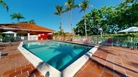 Located at 12375 SW 18th St in Miami, Florida, this outdoor pool area showcases a classic South Florida aesthetic defined by vibrant color, tropical greenery, and open-air leisure. The centerpiece is a generously sized rectangular swimming pool with crystal-clear turquoise water, framed by a terracotta tile deck that adds warmth and a Mediterranean character to the setting. Depth markers are visible along the pool's edges, indicating a gradual transition from shallow to deeper water. Surrounding the pool, a well-appointed deck offers ample space for relaxation, furnished with red lounge chairs and chaise lounges positioned to take full advantage of Miami's abundant sunshine. Shade options are provided by patio umbrellas and a dedicated poolside pavilion featuring a striking red exterior and a clay tile roof, which also serves as a pool house facility. The perimeter of the pool area is enclosed by a white fence and lush, mature tropical landscaping, including towering palm trees and dense canopy trees that provide a sense of privacy and a resort-like ambiance. The multi-story residential building, accented with bold red and neutral tones, rises in the background, complementing the colorful outdoor environment. This amenity space benefits from abundant natural light and open skies, characteristic of Miami's sunny climate.