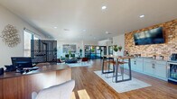 This leasing center in Millington, Tennessee, is a thoughtfully designed single-floor space that balances professional functionality with an inviting, contemporary aesthetic. The open-concept layout is unified by warm hardwood-style flooring and recessed lighting throughout, creating a bright and cohesive environment.

The reception area anchors the space near the entrance, featuring a substantial walnut-toned desk with guest seating positioned across from it, allowing staff to greet visitors comfortably. Natural light flows in through multiple windows and glass-paned entry doors, enhancing the welcoming atmosphere.

A decorative chevron-patterned room divider subtly separates the reception zone from the lounge area, which is furnished with a plush green sofa, accent chairs, and an area rug. Bold abstract artwork lines the walls, and large glass doors open to an outdoor balcony, providing outdoor views and additional natural light.

An exposed brick accent wall anchors the amenity counter, which features light blue cabinetry with granite countertops and a wall-mounted television above. Geometric hanging planters add organic texture to this zone.

A high-top table with bar stools occupies a central collaborative area, while a glass-walled private office is accessible toward the rear. The overall ambiance is modern, polished, and purposefully designed to create a positive first impression.