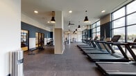 This well-appointed fitness center offers a comprehensive workout environment designed with both functionality and aesthetics in mind. The space is organized into distinct zones, beginning with a dedicated cardio area lined with a row of Star Trac treadmills positioned along expansive floor-to-ceiling windows that flood the room with natural light and provide open outdoor views. Ceiling fans and stylish pendant lights in matte black hang overhead, contributing to a modern, industrial-inspired atmosphere. The flooring transitions between polished concrete in the cardio zone and rubberized flooring in the strength training area, offering practical surfaces suited to each activity. The strength zone is equipped with cable machines, weight benches, and spin bikes, complemented by wall mirrors that enhance the sense of space. Bold navy accent walls adorned with geometric graphic artwork give the fitness center a distinctive visual identity. A glass-door entryway with a hand sanitizer station welcomes users into the facility, while a small display shelf near the entrance adds a thoughtful organizational touch. Ceiling fans ensure comfortable airflow throughout. Natural light pours in from multiple window walls, creating a bright and energizing environment well-suited to an active lifestyle.