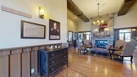 Located at Fort Riley, Kansas, this single-story community space presents a generously sized floor plan unified by a distinct Western frontier aesthetic. The main living area features rich hardwood flooring, soaring ceilings with exposed dark wood beams, and a dramatic floor-to-ceiling stacked stone fireplace that serves as a focal point. Warm-toned upholstered seating, antique-style wood furnishings, and decorative wall art—including historical portraits and Kansas-themed pieces—reinforce the space's regional character throughout.

The hallway transitions between distinct zones, connecting the main lounge to additional gathering areas. One section transitions to plush carpeting and showcases large windows framed by floor-length drapes, allowing substantial natural light and views to the outdoors. This seating area features a pair of classic leather armchairs alongside a central side table, creating an inviting reading or conversation nook.

A dedicated recreation area includes a full-sized billiards table set beneath a period-style chandelier, with vertical wood-paneled walls adorned by vintage Kansas memorabilia, wagon wheel accents, and framed landscape photography. A compact dining table with chairs provides additional seating nearby. Throughout the space, layered lighting—including wall sconces, pendant fixtures, and chandeliers—contributes to a warm, welcoming ambiance with a strong sense of historical identity.