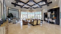 This sophisticated single-floor space makes a striking first impression with its grand entrance foyer, where polished large-format marble tile floors lead to a circular inlay medallion beneath a domed ceiling fitted with a decorative chandelier. Wall sconces and abundant natural light from floor-to-ceiling windows create a warm, inviting atmosphere throughout.

The centerpiece of the space is a sweeping main lounge area defined by a stunning coffered ceiling with bold dark wood beams arranged in a geometric pattern, accented by recessed lighting and a dramatic multi-arm chandelier that anchors the room. An expansive curved wall of floor-to-ceiling windows and glass doors floods the lounge with natural light and offers open views of the outdoor surroundings. The space is furnished with a refined arrangement of sofas, lounge chairs, and accent tables, all set atop polished cream marble flooring that extends seamlessly throughout.

Adjacent to the main lounge is a bar and seating area, and a fully equipped kitchen featuring sleek dark cabinetry, light stone countertops, a full-size stainless steel French door refrigerator with built-in water dispenser, a beverage cooler, and a microwave. The kitchen's practical layout and ample storage make it well-suited for entertaining and events. Together, the spaces combine architectural elegance with functional design, offering a polished and comfortable environment.