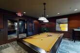 Located at 820 West Marietta St NW in Atlanta's vibrant West Midtown neighborhood, this single-floor amenity space combines industrial character with upscale design. The billiard room is a standout feature, centered around a full-size pool table with elegant curved legs and a gold felt surface, illuminated by pendant lights. Rich mahogany wall paneling, textured carpet, and a pass-through window connecting to an adjacent area give the room a sophisticated club-like atmosphere. The main lounge area exudes a warm, loft-inspired aesthetic, showcasing exposed brick walls, rich hardwood flooring, and dark ceilings with exposed ductwork that add to its industrial appeal. Wrought iron railings border an interior mezzanine overlook, while arched windows flood the space with natural light and offer views of the surroundings. Comfortable seating arrangements, pendant lighting, and a wet bar with counter seating complete the lounge's inviting ambiance. A dedicated business center provides a focused workspace, featuring a mosaic-tiled accent wall, built-in desk stations, and task lighting — designed for productivity within a visually distinctive setting. Together, these spaces offer a well-rounded and thoughtfully designed environment ideal for both socializing and working.