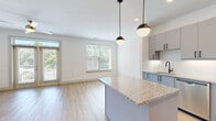 This well-designed 2-bedroom, 1-bathroom residence offers a thoughtfully laid-out single-floor plan with a clean, modern aesthetic. Light wood-look flooring runs throughout the main living areas, complemented by crisp white walls that enhance the bright, airy atmosphere. The open-concept kitchen and living room form the heart of the home, where the kitchen features a granite-topped island with pendant lighting overhead, sleek gray cabinetry with matte black hardware, stainless steel appliances, and a subway tile backsplash. The living area flows naturally from the kitchen, bathed in natural light from multiple windows and a glass door that opens to a private balcony, bringing in lush outdoor views. The primary bedroom is a well-sized retreat with a ceiling fan, large windows, and direct balcony access via glass French doors. The second bedroom offers plush carpeting, a ceiling fan, and a generous window with garden views. The full bathroom is finished with a granite-topped gray vanity, a large framed mirror, and matching hardware consistent with the home's modern design language. A spacious walk-in closet with wire shelving provides ample storage. The overall ambiance is bright, clean, and contemporary throughout.