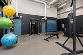Located at 1143 First Ave S in Birmingham, Alabama, this fitness facility is designed to support a comprehensive workout experience within a single-floor layout. The space is characterized by soaring double-height ceilings supported by prominent structural columns, exposed ductwork overhead, and a bold two-tone color scheme of teal and gray that gives the interior a modern, energetic feel.

The main workout floor is generously sized and organized into distinct training zones. A cardio section features multiple treadmills, a rowing machine, and a stationary bike, all positioned beneath wall-mounted flat-screen televisions. Full-length mirrors line one wall, enhancing the sense of openness. A dedicated strength and functional training area includes a dual cable machine, a heavy punching bag, a dumbbell rack, stability balls, medicine balls, resistance bands, and yoga mats.

Large sliding glass doors along one wall open to an outdoor area, filling the space with natural light and providing views of greenery. An interior mezzanine-level railing overlooks the main floor, adding architectural depth. A support area toward the rear of the facility includes gender-designated restrooms, shower facilities, and a bank of blue lockers, offering practical amenities for users before and after training sessions.