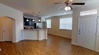 This single-floor space offers a seamless open-concept layout that connects the kitchen and living areas into one cohesive, functional environment. The kitchen is a standout feature, showcasing dark cabinetry that contrasts elegantly with granite countertops and a curved breakfast bar peninsula. Stainless steel appliances, including a refrigerator and built-in microwave, add a modern touch, while three pendant lights suspended above the counter provide both task lighting and visual appeal. A corner shelving unit offers additional storage and display space. The living area flows naturally from the kitchen, highlighted by warm hardwood-style flooring that runs throughout the space, creating a sense of continuity and warmth. Neutral-toned walls keep the atmosphere light and versatile. Natural light enters through multiple windows fitted with white horizontal blinds, brightening the interior without sacrificing privacy. A ceiling fan with an integrated light fixture is centrally positioned in the living area, ensuring comfort and air circulation. Recessed lighting complements the overall illumination scheme. The space presents a clean, contemporary aesthetic that is both practical and welcoming, ideal for everyday living and entertaining alike.