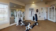 This fitness center offers a thoughtfully designed workout space that balances functionality with an inviting atmosphere. The layout is divided into distinct activity zones, beginning with a strength training area housing multiple white-framed Hoist cable machines and weight stations positioned alongside floor-to-ceiling mirrors that visually expand the space and allow for proper form monitoring during exercise.

The cardio zone is highlighted by a graceful bay window alcove, where curved walls of multi-pane windows flood the area with natural light and provide outdoor views of the surrounding neighborhood. This section accommodates an elliptical trainer, stationary bike, and a Landice treadmill, all arranged to take advantage of the scenic outlook. Additional windows fitted with white horizontal blinds line other walls, ensuring consistent brightness throughout.

Underfoot, durable carpet covers the entire floor, providing cushioning and traction during workouts. Recessed lighting and a ceiling fan contribute to a comfortable, well-ventilated environment. Framed artwork and traditional molding details add a refined aesthetic touch. French doors with glass panels connect to adjacent spaces, enhancing both accessibility and the overall sense of openness within this well-appointed fitness facility.