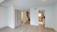 This two-story residence offers a thoughtfully designed layout that balances functionality and comfort across both levels. The main floor showcases light-toned luxury vinyl plank flooring that extends throughout, lending the space a clean, cohesive aesthetic. The open living area is bright and airy, with crisp white walls and a textured ceiling that adds subtle character. A hallway connects key areas of the main floor, including a conveniently located bathroom accessible from the common space. The kitchen, visible from the main living area, features white cabinetry, stainless steel appliances, and a window that invites natural light into the space. A staircase with a classic metal railing leads to the second floor, providing a natural visual anchor to the layout. HVAC vents are integrated into the ceiling, ensuring year-round comfort throughout. The overall ambiance is fresh and move-in ready, with neutral tones and modern fixtures that create a versatile backdrop suited to a variety of interior styles. The two-floor configuration provides a natural separation between living and private spaces, making this a practical and well-organized home.