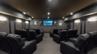This single-level private home theater is thoughtfully designed to deliver an immersive cinematic experience. The room is finished in deep charcoal and dark gray tones throughout, with a dark ceiling that enhances the theater atmosphere and recessed lighting providing controlled, ambient illumination. Rows of plush black leather reclining seats with built-in cup holders are arranged in a tiered, stadium-style layout, ensuring unobstructed sightlines from every position and comfortably accommodating a generous number of viewers. A large wall-mounted flat-screen display serves as the focal point of the screening wall, flanked by framed classic movie posters that add a cinematic character to the space. The side and rear walls are adorned with decorative acoustic panels featuring a geometric patterned fabric, complemented by wall sconces that provide subtle, warm accent lighting. Velvet-style rope stanchions add a charming theater lobby touch near the screen wall. At the rear of the room, double doors open to a concession-style area with shelving and a popcorn machine, elevating the full movie-going experience. The carpeted flooring and sound-absorbing wall treatments contribute to the room's strong acoustic design, making this a well-appointed and fully dedicated entertainment space.