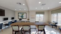 Located on N Schisler Drive in Marana, Arizona, this single-floor community space is thoughtfully designed to serve as a versatile hub for residents. The open-concept layout is anchored by a substantial rustic wood communal table with industrial-style legs, surrounded by patterned upholstered bar-height chairs — an ideal setting for casual gatherings or collaborative work sessions.

Recessed lighting throughout the space ensures bright, even illumination, complemented by natural light streaming through large windows dressed in bold chevron-patterned Roman shades. The windows frame views of the surrounding desert landscape, adding to the relaxed Southwestern ambiance.

A dedicated business center lines one wall, featuring a sleek white built-in desk with multiple computer workstations and a large wall-mounted flat-screen television above. Decorative metalwork wall sculptures add artistic interest throughout the space.

A welcoming lounge nook near the front windows offers comfortable navy upholstered chairs with accent pillows, positioned to take in the outdoor views — perfect for reading or informal conversation. Light-toned flooring and neutral warm walls tie the space together, creating a clean, contemporary aesthetic. An open passageway connects seamlessly to adjacent areas, enhancing the overall flow and accessibility of this well-appointed community space.