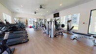 This modern fitness center offers a spacious, single-floor layout designed to support a full range of workout routines. The interior features warm wood-look flooring throughout, crisp white walls, and recessed lighting complemented by dual ceiling fans that promote air circulation during workouts. Large windows line multiple walls, flooding the space with natural light and offering views of lush outdoor greenery and the surrounding property.

The cardio zone is well-stocked with a row of True-brand treadmills and upright stationary bikes, positioned to take advantage of the natural light and outdoor views. A dedicated strength training area houses a multi-station cable and weight machine, a Smith machine with an adjustable bench, and additional resistance equipment for targeted muscle training. Full-length mirrors are strategically placed to enhance the sense of space and allow users to monitor their form.

A free weights section provides a complete dumbbell rack with a wide range of weights and an adjustable workout bench, accompanied by stability and medicine balls for functional training. A wall-mounted flat-screen television adds an entertainment element to the training environment. The space is accessed via a glass-paneled entry door that opens to the outdoors, and includes a restroom or utility area accessible from within. Overall, the fitness center delivers a clean, bright, and professional environment suited for comprehensive daily training.