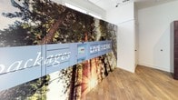The package room is a well-appointed, single-floor amenity space designed to streamline parcel delivery and retrieval for residents. The room is defined by two distinct locker systems: a set of dark espresso-finished package lockers with multiple compartment sizes lining one wall, and a bank of smart package lockers integrated into a striking floor-to-ceiling nature-themed mural on the opposite wall. The mural, depicting towering trees bathed in golden sunlight, is branded with "Live Here" and "Secure Packages" signage, adding a visually dynamic element to an otherwise functional space. The smart locker system features a touchscreen interface, enabling secure, self-service package pickup. Wood-style plank flooring in a warm, natural tone runs throughout, complementing the clean white walls and angled ceiling. Natural light filters into the room through multiple windows, while a direct exterior door provides convenient access from outside. A security camera is mounted near the ceiling, ensuring a monitored and secure environment. The overall design balances practicality with thoughtful aesthetic touches, creating a functional yet welcoming package management space.