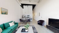 This contemporary loft-style residence offers two bedrooms and two bathrooms across a single floor, combining industrial architectural elements with modern finishes. The open-concept living, dining, and kitchen area is anchored by dramatically high ceilings with exposed ductwork and pendant lighting, creating an airy, urban atmosphere. Polished concrete floors run throughout the main living space, while floor-to-ceiling glass entry doors flood the area with natural light and provide access to an outdoor walkway.

The kitchen is thoughtfully designed with two-tone cabinetry — grey lower cabinets paired with crisp white uppers — white quartz countertops, a subway tile backsplash, and a full suite of stainless steel appliances including a gas range, microwave, and French door refrigerator. A peninsula counter doubles as a dining bar adjacent to the dining area.

The primary bedroom is generously proportioned with soaring ceilings and soft carpeting, offering a calm retreat. The second bedroom mirrors this serene aesthetic with tall white walls and plush carpet. Both bathrooms are finished in clean, modern white, with one featuring a double-sink vanity with white countertops and a large mirror, and the other showcasing a deep soaking tub surrounded by large-format tile.