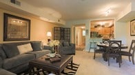 This two-floor residence offers a thoughtfully designed layout that balances comfort and functionality. The main living area is open and inviting, featuring plush carpeted flooring, warm neutral wall tones, and ample space for both relaxing and entertaining. Comfortable upholstered seating surrounds a classic leather ottoman coffee table, while a decorative geometric wall panel adds a distinctive artistic touch. Soft ambient lighting from a table lamp and overhead fixtures creates a cozy atmosphere throughout.

The living space flows seamlessly into a dedicated dining area, where a round dark-wood table is paired with cushioned chairs, providing an ideal setting for meals. Just beyond, the kitchen is visible through a pass-through breakfast bar with counter seating, offering a practical and social connection between cooking and dining spaces. The kitchen features warm wood cabinetry, a chandelier-style light fixture, and efficient counter space.

A hallway visible from the main living area leads further into the residence, hinting at additional rooms across both floors. Natural light filters in through windows, brightening the interior and complementing the warm, earthy color palette. Overall, the space presents a welcoming and well-organized home environment suited for everyday living.