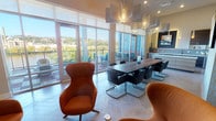 This elegantly designed single-floor amenity space masterfully blends functionality with upscale aesthetics. The open layout is anchored by a wall of floor-to-ceiling glass doors that flood the interior with natural light and frame stunning panoramic views of a waterway and lush treeline beyond a furnished outdoor terrace.

The main gathering area features a long, solid-wood conference table surrounded by contemporary black cantilever chairs, ideal for meetings or group events. Statement pendant lighting with sculptural, geometric fixtures hangs overhead, complementing the herringbone-patterned tile flooring that flows throughout the space.

A dedicated lounge nook offers a more intimate setting, furnished with bold burnt-orange egg-style chairs arranged around a low wood cube table, all centered on a large flat-screen television set into a sleek, wood-paneled accent wall. Curated artwork and a large mirror add depth and character to this area.

A well-appointed kitchenette lines one wall, complete with dark wood-grain cabinetry, white quartz countertops, a tile backsplash, stainless steel appliances, and under-cabinet accent lighting. A bold abstract painting serves as a dramatic focal point above the cabinetry. Built-in open shelving along another wall provides stylish display space, completing this refined and versatile amenity suite.