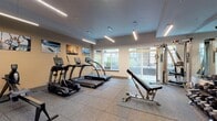 This fitness center is a thoughtfully designed, single-floor facility built to accommodate a full range of workout routines. The space features durable rubber flooring throughout, providing both safety and comfort during exercise. Overhead, sleek linear pendant lighting illuminates the room with a clean, modern aesthetic, complemented by natural light streaming in through large windows and glass doors that open to an outdoor balcony area.

The cardio zone is anchored by a row of TRUE-brand treadmills and elliptical trainers, offering users professional-grade equipment for aerobic training. A rowing machine rounds out the cardio selection. For strength training, the space includes a multi-station cable machine, a full rack of dumbbells spanning a wide range of weights, an adjustable workout bench, and a stability ball. A floor-to-ceiling mirror along one wall visually expands the space and allows users to monitor their form during exercises.

The warm tan accent wall is decorated with a series of large-format outdoor and adventure-themed photographs, lending an energizing and motivational atmosphere to the room. A water fountain and a small service counter are conveniently located near the entrance. The overall design balances functionality with a polished, welcoming environment suited for a variety of fitness levels and training styles.