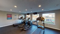 Located at 507 Stone Creek Way in Cincinnati, Ohio, this single-level fitness facility is designed to support a comprehensive workout experience across multiple interconnected zones. The space is finished with durable dark rubber flooring throughout, providing a practical and resilient surface suited for a wide range of exercise activities.

The cardio area houses multiple treadmills and elliptical machines arranged in rows, with large windows along one wall allowing ample natural light and views of the surrounding outdoor area. Wall-mounted televisions are positioned for convenient viewing during cardio sessions. Recessed lighting and a textured ceiling contribute to a clean, functional aesthetic throughout.

A dedicated strength training zone features a multi-station cable machine, an adjustable weight bench, a dumbbell rack with free weights, and a large wall mirror that enhances the sense of space and assists with exercise form. Instructional fitness posters are displayed on the walls throughout, offering workout and stretching guidance.

Additional areas include open floor space for functional training, a medicine ball rack, a balance board, and a folding mat station. Built-in shelving and a small work counter provide organizational convenience, while a defibrillator unit is mounted on the wall, reflecting a safety-conscious design. The facility also includes clearly marked exit access.