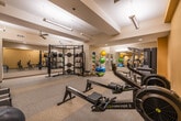 This fitness center offers a thorough and well-organized workout environment designed to accommodate a wide variety of exercise routines. The space is finished with warm golden-yellow walls and durable speckled flooring, creating an energetic yet comfortable atmosphere. Recessed lighting and overhead fluorescent fixtures provide consistent, even illumination throughout, while ceiling fans promote air circulation across the main floor.

The cardio section is well-appointed with multiple treadmills, rowing machines, elliptical trainers, and a stationary bike, offering ample options for aerobic training. Strength training is equally well-represented, with a multi-station cable machine, a power rack stocked with weight plates, barbells, and kettlebells, as well as a full dumbbell rack. A dedicated free weights area includes an adjustable bench and a broad selection of dumbbells for targeted resistance work.

Functional training is supported by an extensive collection of medicine balls and stability balls displayed on a dedicated rack. Full-length mirrors line one wall, enhancing the sense of openness and allowing users to monitor their form during workouts. The overall layout is thoughtfully arranged to ensure sufficient space between equipment, providing a safe and efficient training environment suitable for users of all fitness levels.