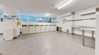 This shared laundry facility, located in Toronto, Ontario, offers a generously sized, single-floor space designed for efficient and comfortable use. The room is finished with light grey large-format floor tiles throughout, contributing to a clean and bright aesthetic. White walls are accented by a horizontal tan tile border that runs along the perimeter, adding a subtle decorative touch.

The facility is equipped with a substantial lineup of front-load and top-load washers and dryers, arranged along the walls to maximize open floor space in the centre. Fluorescent strip lighting runs across the ceiling, ensuring the room is well-lit at all times. A row of high, frosted horizontal windows allows natural light to filter in while maintaining privacy.

A dedicated folding and sorting area is provided via built-in stainless steel countertops along one wall, offering practical workspace for residents. A bench is available for those waiting, adding a comfortable touch to the space. Informational signage, a bulletin board, and safety equipment such as a fire extinguisher are clearly visible and accessible. A utility sink and waste disposal station are also present, rounding out the room's functionality.

The layout is open and uncluttered, making it easy to navigate, and the overall ambiance is clean, orderly, and efficiently designed for shared residential use.
