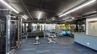 This fitness facility offers a comprehensive workout environment designed to accommodate a wide range of exercise needs. The space features an industrial aesthetic, with an exposed ceiling showcasing ductwork, piping, and bright linear fluorescent lighting that ensures excellent visibility throughout the floor.

The main workout area is generously sized and thoughtfully organized. A full dumbbell rack lines one side, offering an extensive range of free weights for strength training, accompanied by adjustable benches positioned nearby for versatile exercises. Cardio machines, including upright bikes and ellipticals, are visible toward the rear of the space.

Functional training is well supported by a dedicated area stocked with stability balls in multiple sizes, medicine balls on tiered racks, balance discs, and resistance accessories. Rubber flooring tiles are laid over blue-toned carpet, providing both safety and comfort underfoot.

Full-length mirrors line the walls, enhancing the sense of depth and allowing users to monitor their form during workouts. The open layout promotes easy movement between equipment zones, creating a practical and motivating environment suited for individual training sessions.
