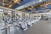 Located at 2315 South 11th Avenue in Bozeman, Montana, this fitness center offers a well-appointed, single-floor workout facility designed to meet a wide range of exercise needs. The space features an open, industrial-style interior with an exposed ceiling showcasing ductwork, structural elements, and bold navy blue accent walls adorned with motivational lettering — creating an energetic and focused atmosphere throughout.

Large floor-to-ceiling windows line the perimeter walls, flooding the space with natural light and providing views of the surrounding Bozeman landscape. Oversized ceiling fans and suspended linear lighting ensure consistent airflow and visibility across the entire floor.

The main workout area is organized into distinct training zones. A dedicated cardio section houses a generous row of treadmills, elliptical machines, and stair climbers, all oriented toward the windows. The free weights area features a full dumbbell rack, kettlebells, adjustable benches, and barbells, complemented by full-length mirrors for form monitoring. A strength training zone includes a squat rack and cable machines suitable for a variety of resistance exercises.

Mounted televisions and motivational wall graphics enhance the workout environment. Separate restroom facilities for ladies and gents are clearly marked. The durable rubber flooring throughout supports safe and comfortable training across all activity zones.
