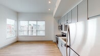 This single-floor unit features a contemporary, open-concept layout that seamlessly connects the kitchen and living area, creating a bright and functional living space. Light hardwood flooring extends throughout, lending warmth and visual continuity across the unit.

The kitchen is well-appointed with stainless steel appliances, including a range with an electric cooktop, built-in microwave, refrigerator, and dishwasher. Gray flat-front cabinetry lines the walls, providing ample upper and lower storage, while a white countertop offers generous workspace. A white subway tile backsplash adds a clean, modern accent. A single-basin sink with a contemporary faucet completes the kitchen setup.

The open living area benefits from a large window fitted with horizontal blinds, allowing natural light to fill the space while maintaining privacy. Recessed lighting throughout provides consistent overhead illumination.

The bathroom includes a double-sink vanity topped with a white countertop, a large wall mirror, grab bars for accessibility, a toilet, and a glass-enclosed walk-in shower. The hardwood-style flooring continues into this space.

A dedicated laundry closet houses a stacked Whirlpool washer and dryer unit, adding everyday convenience. Additional doors visible in the unit suggest access to a bedroom and closet space.