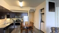 Located at 1301 Washington St in historic Vicksburg, Mississippi, this single-floor residence offers a functional and thoughtfully designed living space. The kitchen is outfitted with stainless steel appliances, including a refrigerator, electric range, and over-the-range microwave, all complemented by rich dark cabinetry and a tile backsplash. Dark hardwood-style flooring runs throughout, lending a warm, cohesive aesthetic to the interior. A dedicated dining area sits adjacent to the kitchen, providing a comfortable space for everyday meals, and is served by a wall-mounted mini-split climate control unit for efficient heating and cooling. A notable convenience of this unit is the in-unit laundry closet, which houses a stacked washer and dryer, adding a practical touch to daily living. The layout is efficient and well-organized, with a clear flow between the kitchen, dining, and utility areas. The space is illuminated by overhead lighting, creating a bright and welcoming interior atmosphere. Overall, this unit delivers a blend of modern amenities and practical design in a convenient Vicksburg location.