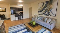 This single-floor residence showcases an open-concept floor plan that integrates the living room and kitchen into one cohesive, functional space. The living area is anchored by a comfortable gray sofa adorned with patterned throw pillows and complemented by a rustic industrial-style coffee table set atop a bold blue-and-white striped area rug. A large abstract painting in blue and gold tones serves as a striking focal point on the main wall, adding a vibrant artistic touch to the space.

The kitchen, visible from the living area, features dark cabinetry, stainless steel appliances including a refrigerator and microwave, and a breakfast bar with seating for two, making it ideal for casual dining and entertaining. The hardwood-style flooring runs consistently throughout both areas, creating a unified and clean aesthetic.

The space is bathed in warm, artificial lighting from ceiling fixtures, contributing to an inviting atmosphere. A neutral wall palette allows the colorful décor and dark cabinetry to stand out. The overall design balances modern and rustic elements, resulting in a stylish yet approachable living environment.