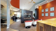 This thoughtfully designed single-floor communal space makes a bold first impression with its striking color palette of burnt orange, deep charcoal, and cool gray. The main lounge area features soaring vaulted ceilings with ceiling fans, dramatic dark wood built-in cabinetry, and a sleek linear fireplace framed by mosaic tile detailing — serving as the visual centerpiece of the room. Modern swivel chairs in white and red provide stylish seating throughout.

A secondary lounge area is warmed by arched transom windows that flood the space with natural light, offering views of an outdoor pool area. Comfortable sectional seating and accent lamps create a relaxed, inviting atmosphere, complemented by patterned carpet flooring and curated wall art.

The fully equipped kitchen features dark espresso cabinetry, stainless steel appliances, a mosaic tile backsplash, and a breakfast bar with bar-height seating. Pendant lighting in amber tones adds warmth and dimension. A round dining table with modern chairs provides additional seating nearby.

A high-bar seating area adjacent to tall arched windows and bold orange accent walls rounds out the space, offering a casual spot for gathering. Throughout, the design strikes a balance between energetic color and refined contemporary finishes, creating a welcoming and functional environment.