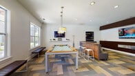 This single-level game room is thoughtfully designed to offer a dynamic blend of recreation and relaxation. At the heart of the space sits a full-size pool table topped with a light felt surface, illuminated by elegant hanging drum pendant lights that draw the eye upward to the clean white ceiling outfitted with recessed lighting. The patchwork-style carpet, featuring a mosaic of blues, greens, tans, and grays, anchors the room with a bold and contemporary aesthetic.

Large windows along one wall flood the space with natural light and offer views of an outdoor pool area, adding to the open, airy feel of the room. A built-in wooden bar counter with white bar stools provides a casual gathering spot, while multiple distinct seating zones cater to different preferences. One area features a generous L-shaped gray sectional sofa with colorful accent pillows and an ottoman, positioned near a wall-mounted flat-screen TV set against a rich dark wood accent wall. A separate nook offers a pair of deep blue armchairs beneath a striking large-scale yellow abstract painting.

Throughout the room, bold abstract artwork punctuates the white walls, adding energy and personality. The overall ambiance is modern, welcoming, and well-suited for social interaction and leisure.