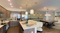 Located at 1109 Mateo Dr in Wilmington, NC, this single-floor community space is designed with both functionality and comfort in mind. The open-concept layout seamlessly connects a well-appointed kitchen area with a social lounge, creating an inviting environment for gathering and relaxation.

The kitchen features a large central island topped with light quartz countertops and equipped with a sink, surrounded by cushioned white bar-height seating. Dark wood cabinetry lines the walls, complemented by a stainless steel refrigerator and a subway tile backsplash. Pendant lighting with decorative patterned shades hangs above the island, adding a touch of elegance to the space.

Adjacent to the kitchen, the lounge area is bathed in warm ambient lighting from wall-mounted sconces, creating a relaxed and welcoming atmosphere. A billiards table serves as a central recreational feature, making the space ideal for socializing and entertainment. The soft sage-green walls and wood-look tile flooring throughout unify the space with a cohesive, modern aesthetic.

Recessed ceiling lighting provides bright, even illumination across the entire floor plan, enhancing both the practicality and visual appeal of this thoughtfully designed communal space.