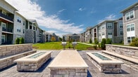 Located at 716 Lemon Dr in Lake Saint Louis, Missouri, this outdoor amenity courtyard is a well-appointed gathering space nestled within a multi-building residential community. The centerpiece of the area is a stamped concrete patio finished in a sophisticated gray tone, anchored by two substantial stone-clad fire pit structures. These fire pits feature glass-fire inserts and wide ledgestone caps, providing both a visual focal point and a functional gathering spot for residents. Adjacent to the fire pits, built-in stone seating walls with matching masonry craftsmanship extend the outdoor living experience. Stainless steel grilling stations are positioned throughout the green space, offering convenient options for outdoor cooking and entertaining. A sweeping, well-maintained lawn stretches between the surrounding multi-story residential buildings, which feature a contemporary design combining board-and-batten siding, stone accents, and private balconies. Thoughtfully placed evergreen shrubs and ornamental trees add greenery and definition to the walkways and patio edges. String lights are strung between poles across the lawn, creating an inviting ambiance for evening use. Paved pedestrian pathways connect the various areas of the courtyard, making it easily accessible. The space benefits from abundant natural light and open sky views, creating a welcoming outdoor environment for the community.