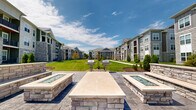 Located at 716 Lemon Dr in Lake Saint Louis, Missouri, this outdoor amenity courtyard is a well-appointed gathering space nestled within a multi-building residential community. The centerpiece of the area is a stamped concrete patio finished in a sophisticated gray tone, anchored by two substantial stone-clad fire pit structures. These fire pits feature glass-fire inserts and wide ledgestone caps, providing both a visual focal point and a functional gathering spot for residents. Adjacent to the fire pits, built-in stone seating walls with matching masonry craftsmanship extend the outdoor living experience. Stainless steel grilling stations are positioned throughout the green space, offering convenient options for outdoor cooking and entertaining. A sweeping, well-maintained lawn stretches between the surrounding multi-story residential buildings, which feature a contemporary design combining board-and-batten siding, stone accents, and private balconies. Thoughtfully placed evergreen shrubs and ornamental trees add greenery and definition to the walkways and patio edges. String lights are strung between poles across the lawn, creating an inviting ambiance for evening use. Paved pedestrian pathways connect the various areas of the courtyard, making it easily accessible. The space benefits from abundant natural light and open sky views, creating a welcoming outdoor environment for the community.