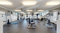 This fitness center offers a thoughtfully designed, single-floor layout divided into distinct functional zones, providing a comprehensive environment for a wide range of workout routines. The main exercise area features durable rubber flooring throughout, providing both safety and comfort underfoot, and is illuminated by a combination of large flush-mount light fixtures and multiple windows fitted with slatted blinds that allow generous natural light to filter in.

The strength training zone is well-appointed with a variety of cable machines, weight benches, a multi-station functional trainer, free weight racks stocked with dumbbells, and plate-loaded bar systems. Mirrored wall sections enhance the sense of space while supporting proper form during exercises. The cardio area is equipped with multiple treadmills, an elliptical trainer, and a stationary bike, alongside a wall-mounted television for entertainment during workouts.

Overhead, multiple modern ceiling fans with polished metal finishes help circulate air throughout the space. Crown molding and white wainscoting add a refined aesthetic detail, complementing the two-tone gray and white wall treatment. A welcoming entryway with wood-look flooring, glass-paneled French doors, and a thermostat control provides a transitional space at the entrance.
